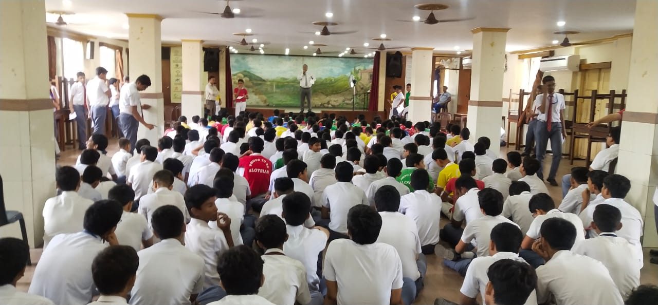 Boys seated on the floor of a hall while a speaker addresses them from the stage