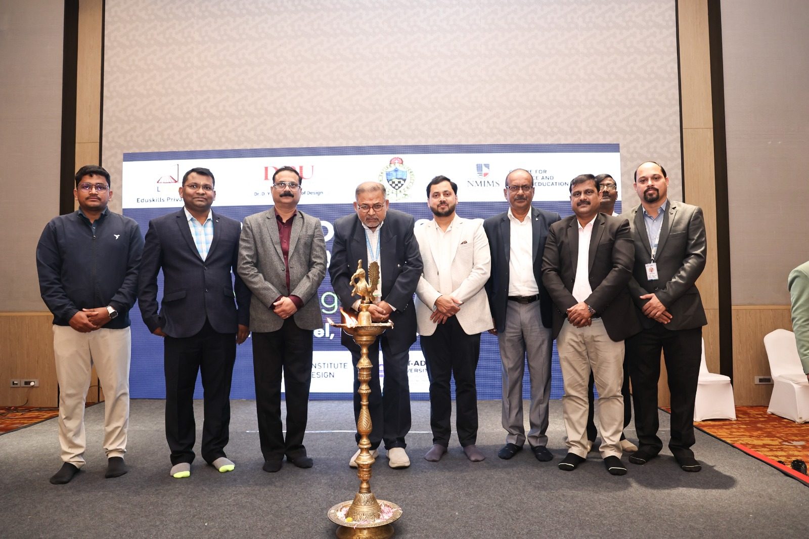 Eight men standing around a ceremonial lamp at an event with partner university banners behind