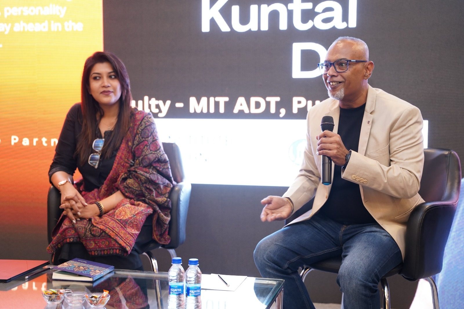 Two panel speakers in conversation on stage, one holding a microphone
