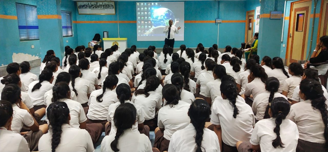 Speaker presenting on a projector screen to girl students seated on the floor